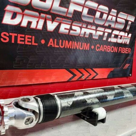 Carbon fiber driveshaft with polished aluminum yoke and performance U-joints, displayed against a red and black background.