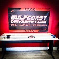 Aluminum driveshaft for 2015+ Dodge Challenger models, featuring a sleek silver finish and performance U-joints, displayed on a black table against a vibrant red background with a promotional banner.