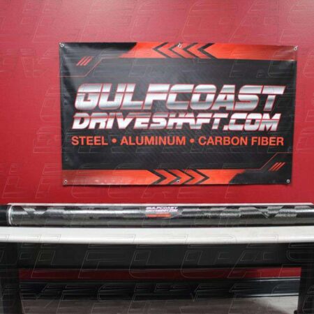 Black and red carbon fiber driveshaft displayed on a counter, with a backdrop featuring "Gulf Coast Driveshaft" branding and a warning.