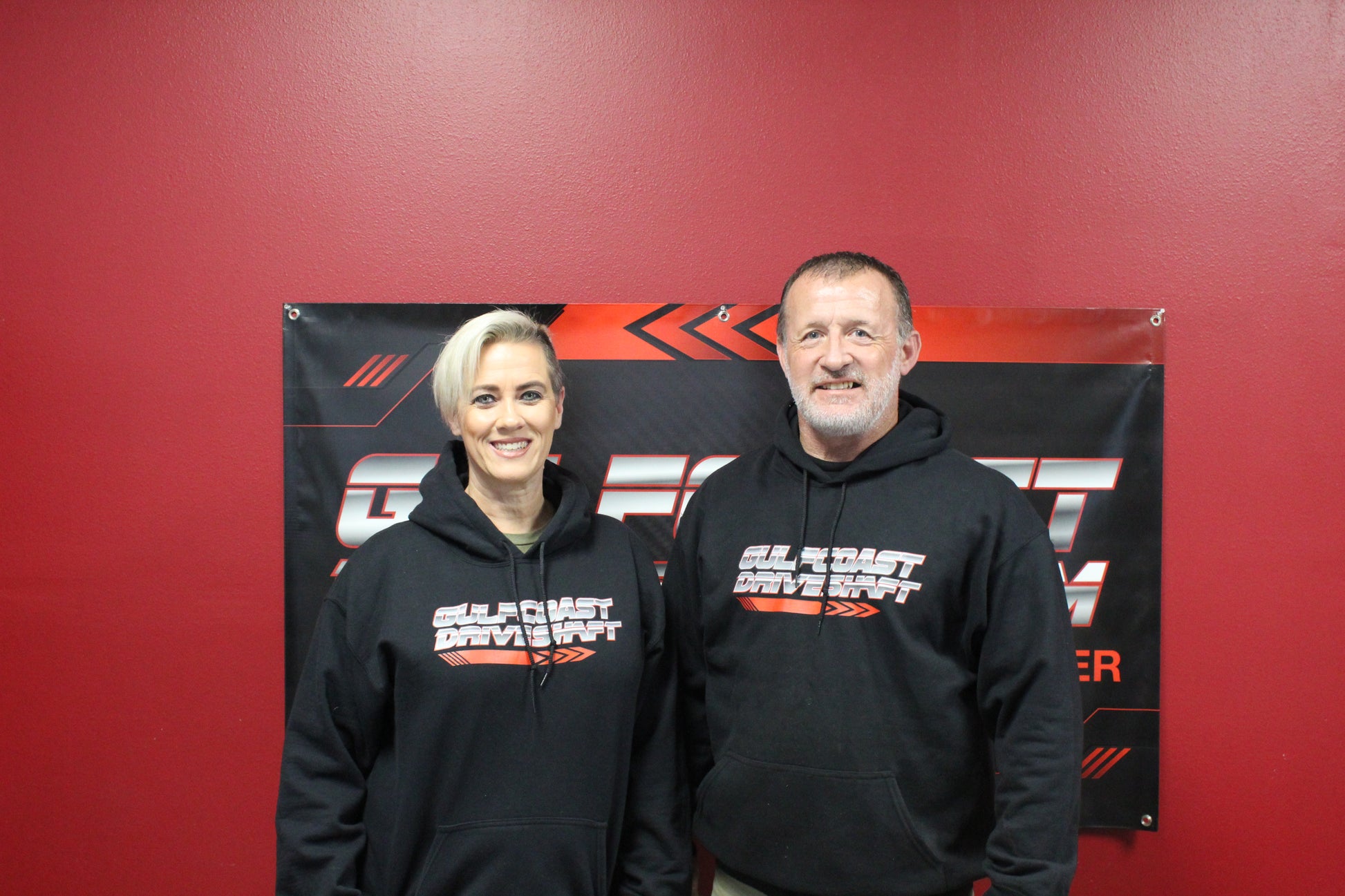 Two people wear black Gulf Coast Driveshaft logo hoodies, showcasing the design against a red background.